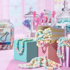Pastel candy necklaces draped over colorful gift boxes and scattered on a pink surface, with a jar of similar candy beads and blurred candy displays in the background.