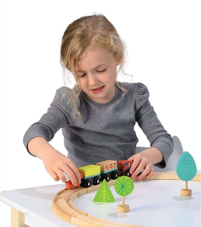 A young child playing with the My First Wooden Train Set by Mentari, assembling the wooden tracks and setting up a toy train, house, and trees on a white surface.