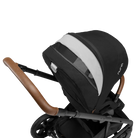 A close-up profile view of the black MIXX Next + Pipa Aire RX Travel System by Nuna stroller showing the brown leatherette handlebar and an extended canopy with an open white ventilation panel, set against a checkered transparent background.