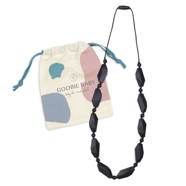 A black beaded silicone teether necklace with a black cord, displayed next to its cream-colored drawstring pouch featuring pastel blue, pink, and gray abstract shapes and the brand name 'GOOBIE BABY'.