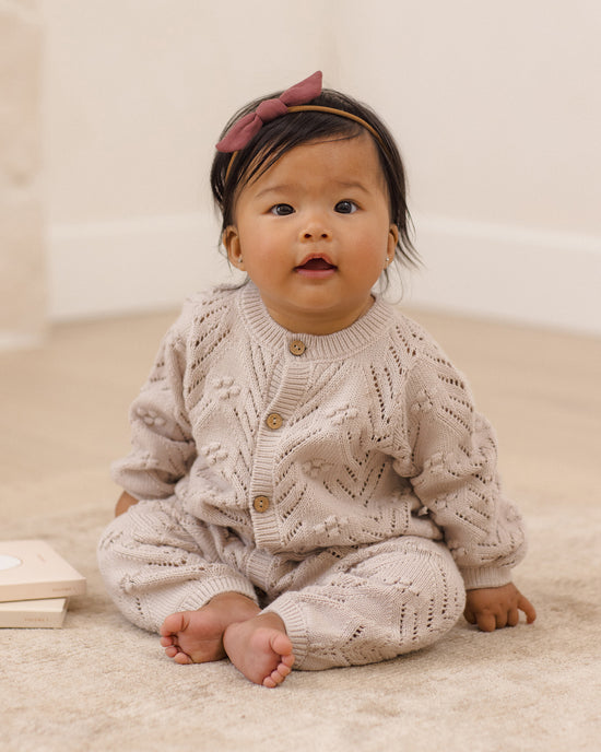 A baby sitting on a beige carpet wearing a matching knit cardigan and pants set in a warm cream-beige color, with a dusty rose bow headband. The background is a soft, neutral indoor room.