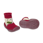 A pair of baby booties with red rubber soles, featuring a cream knit upper with a red cuff and a green apple graphic with the text 'EAT'.
