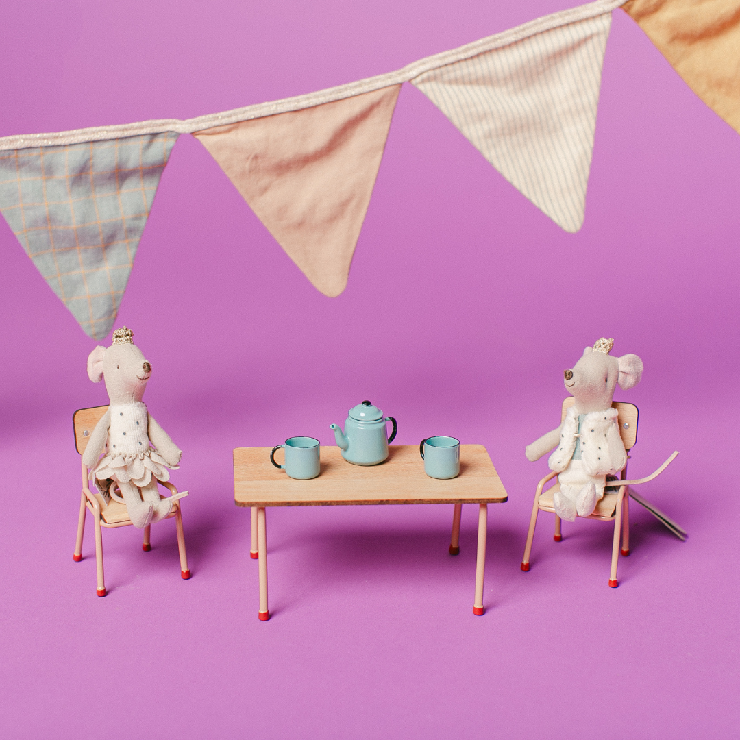 Two stuffed animals sitting at a small table with teacups and a teapot, under a string of flags on a purple background.