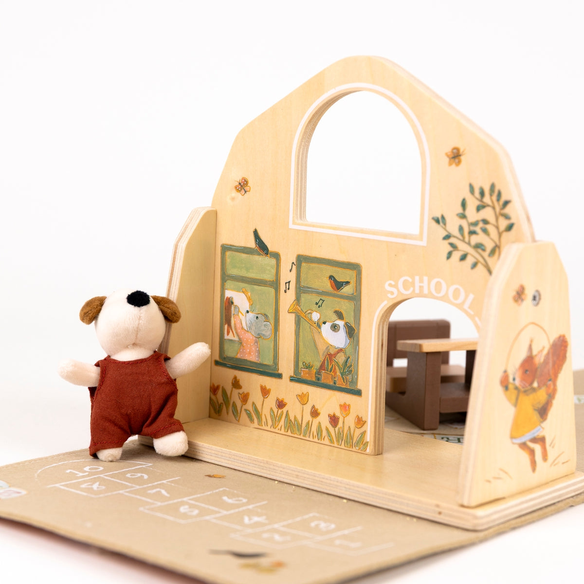 A wooden toy school house with painted animal characters and a small plush dog figure in a red outfit standing beside it on a light neutral background.
