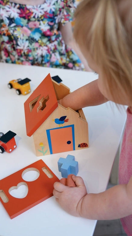 A toddler sits on the floor, holding a blue wooden puzzle piece while exploring a wooden shape sorter house and various colorful geometric blocks scattered nearby.