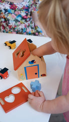 A toddler sits on the floor, holding a blue wooden puzzle piece while exploring a wooden shape sorter house and various colorful geometric blocks scattered nearby.