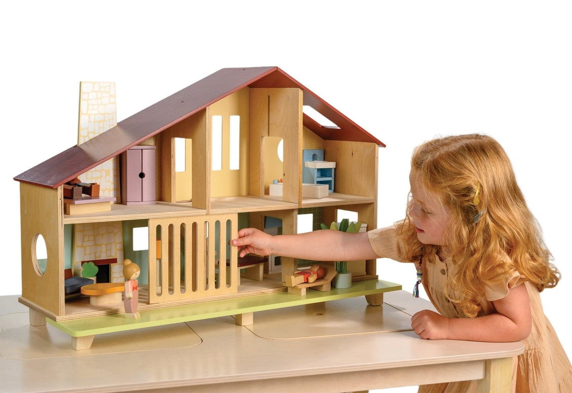 A young child with curly hair and a yellow dress plays with the Wooden Cedar Chalet Modernist Dollhouse by Mentari, sliding open the ground-floor grill on the mid-century modern home while arranging miniature furniture and dolls inside and on the patio.