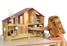 A young child with curly hair and a yellow dress plays with the Wooden Cedar Chalet Modernist Dollhouse by Mentari, sliding open the ground-floor grill on the mid-century modern home while arranging miniature furniture and dolls inside and on the patio.