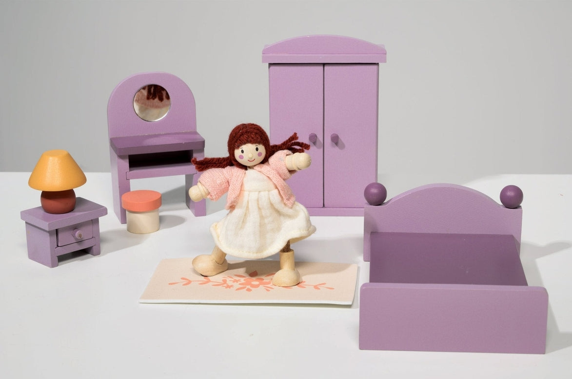 A dollhouse bedroom scene featuring a wooden doll in a pale dress holding a small bag, standing near the center in front of a purple double bed. In the background, a matching purple wardrobe stands beside a dressing table with an oval mirror and a matching stool, while a bedside table with an orange base and yellow shade sits to the left.