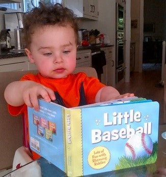 Child holding a book titled 'Little Baseball' in a kitchen setting