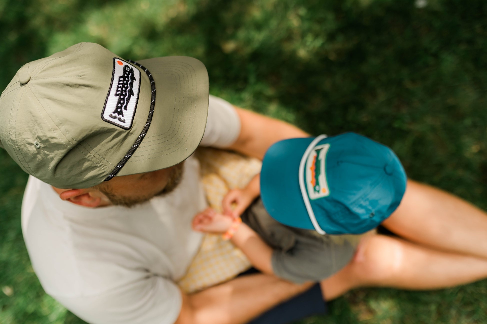 A man and a child sitting together outdoors on grass, both wearing matching khaki and teal fishing caps with a visible 'Fishing Rod' brand patch on the side.