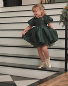 A young girl with blonde hair wearing a dark green puff-sleeve tulle dress and metallic gold shoes, posing on a black-and-white staircase.