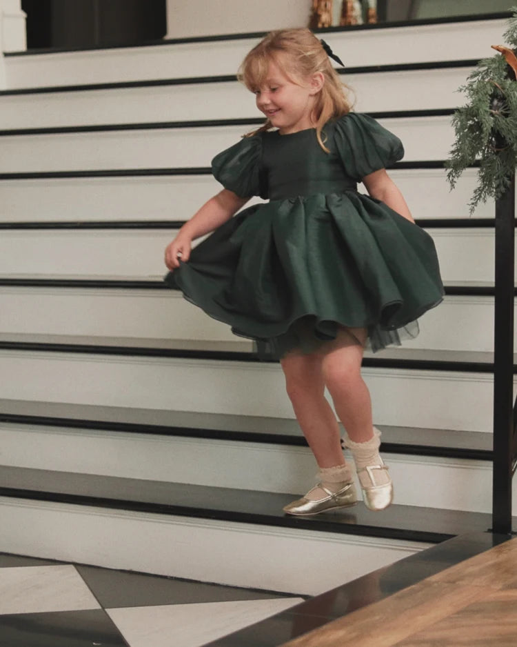 A young girl with blonde hair wearing a dark green puff-sleeve tulle dress and metallic gold shoes, posing on a black-and-white staircase.