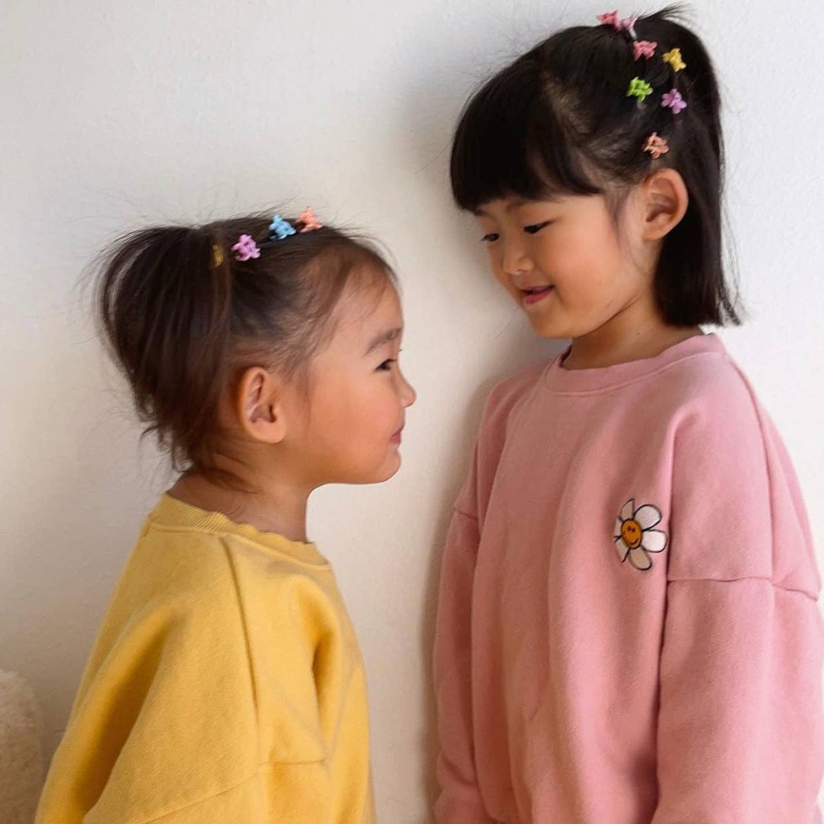Two young girls wearing colorful hair clips and standing close to each other against a plain background.