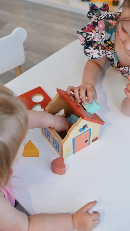 A young child looks down intently as they play with a wooden shape sorter house on a table, with an adult or older child leaning in beside them to watch. Colorful wooden blocks are scattered on the surface near the toy.