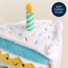 Plush toy shaped like a birthday cake with a candle and sprinkles on a beige background.