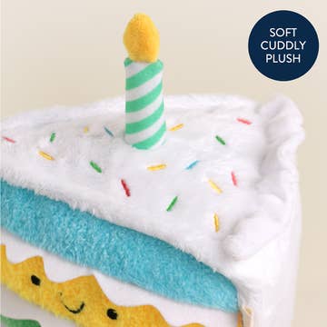 Plush toy shaped like a birthday cake with a candle and sprinkles on a beige background.