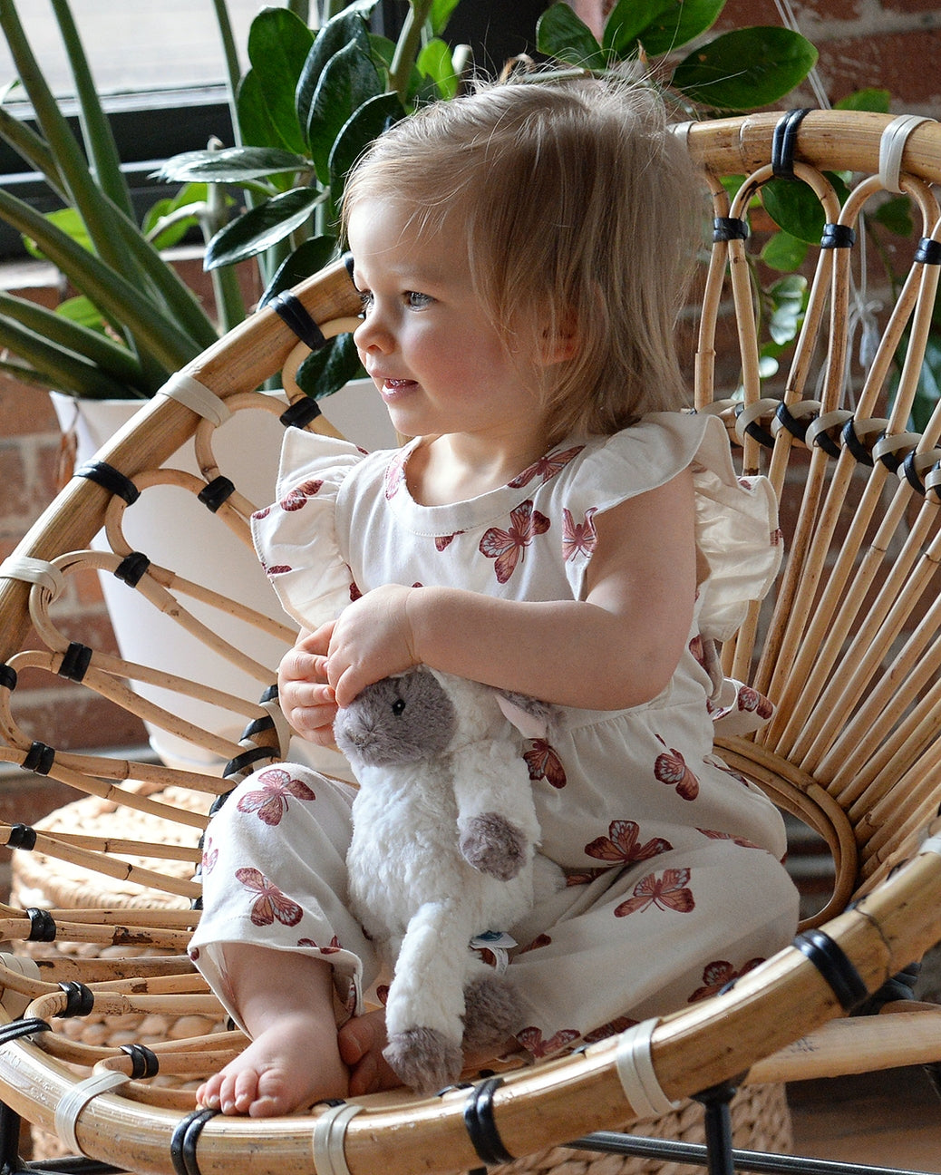Child sitting in a wicker chair holding a stuffed animal, surrounded by plants.