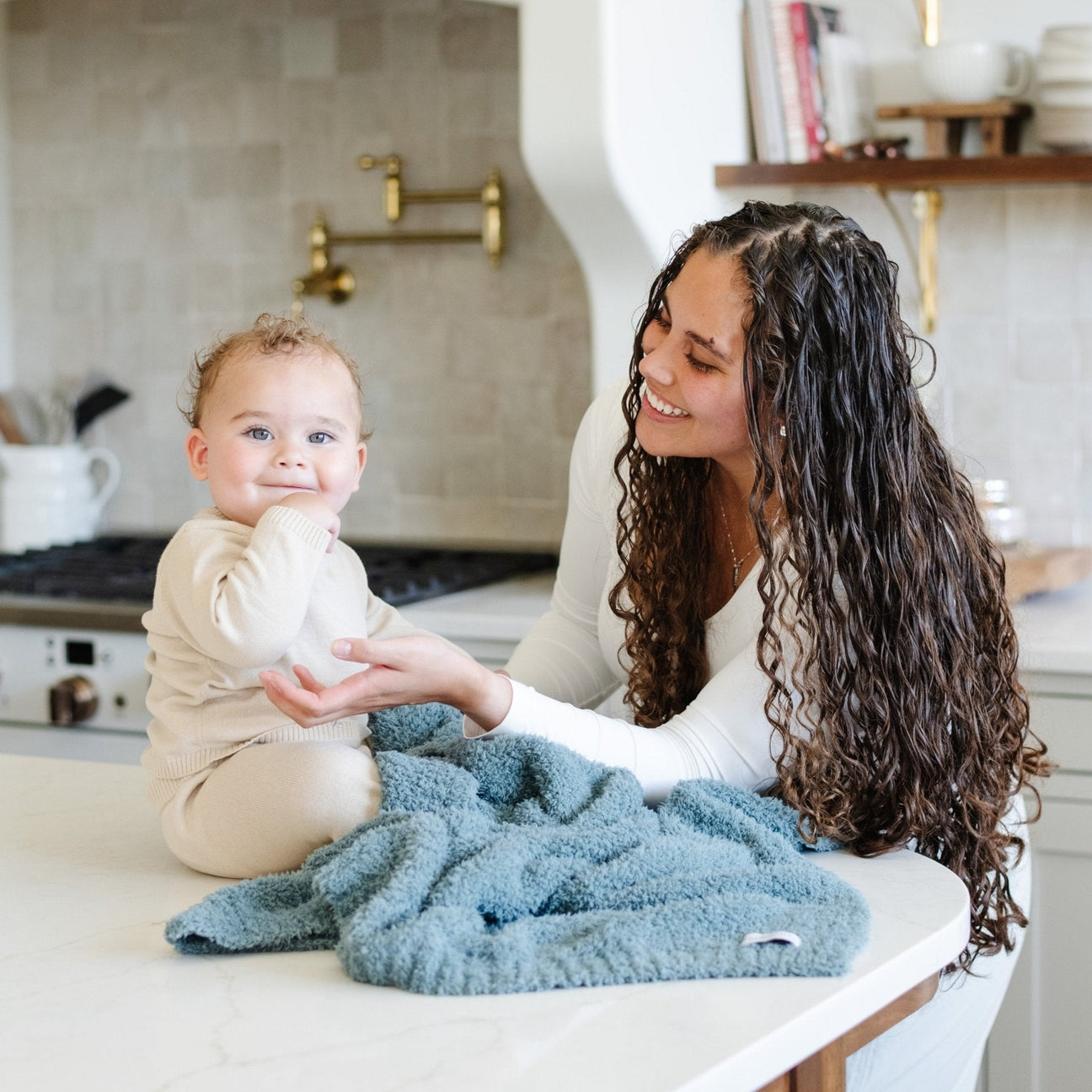 A woman with long curly brown hair smiles and holds a baby sitting on a light blue fleece blanket in a bright, modern kitchen. The baby is wearing a cream-colored outfit.