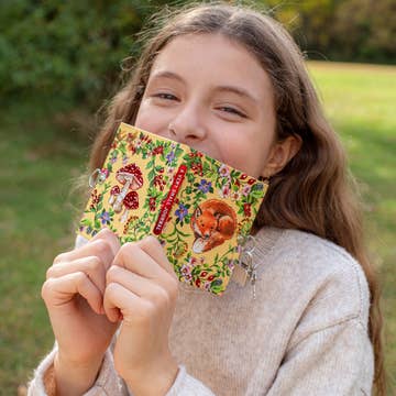 Young girl holding a colorful book with nature-themed illustrations in an outdoor setting.