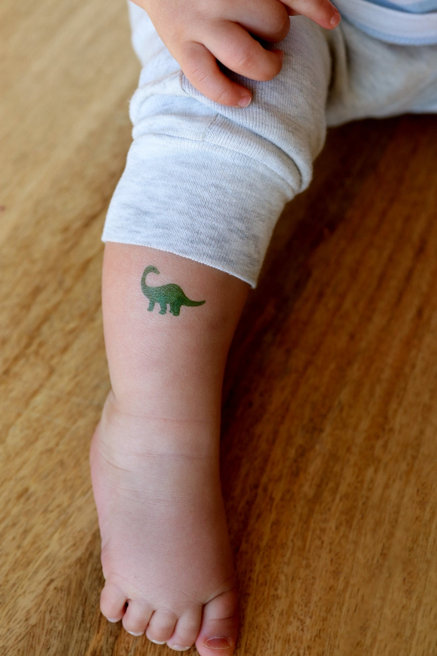Child's leg with a small green dinosaur tattoo on a wooden floor.