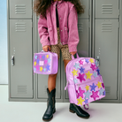 A child stands in front of gray school lockers holding a pink backpack and matching lunch bag. The backpack and lunch box are decorated with colorful star appliqués in pink, purple, blue, and yellow.