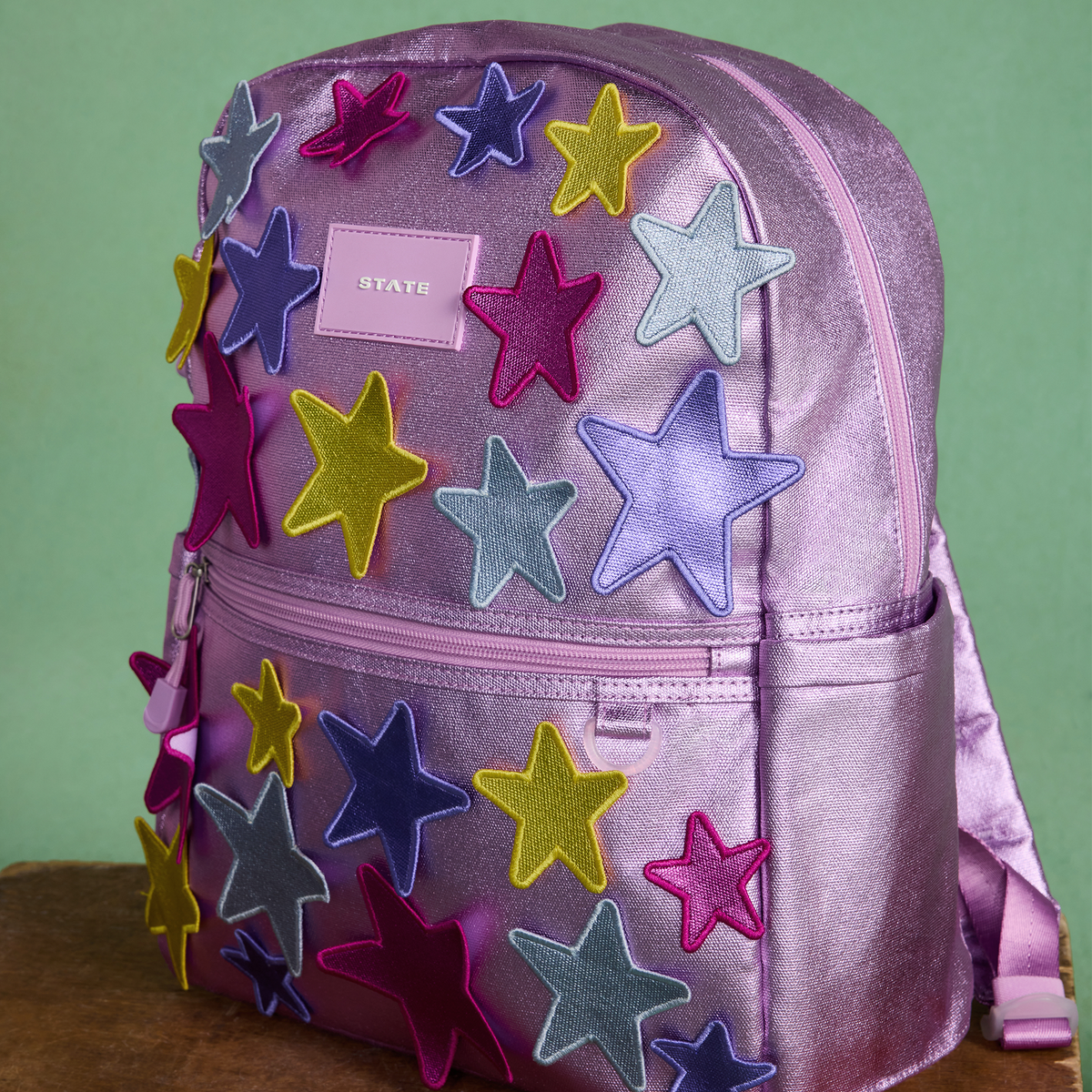 A metallic lavender-purple backpack decorated with multiple colorful felt star appliqués in pink, purple, blue, yellow, and gray, featuring a front zippered pocket and a small pink 'STATE' brand patch.