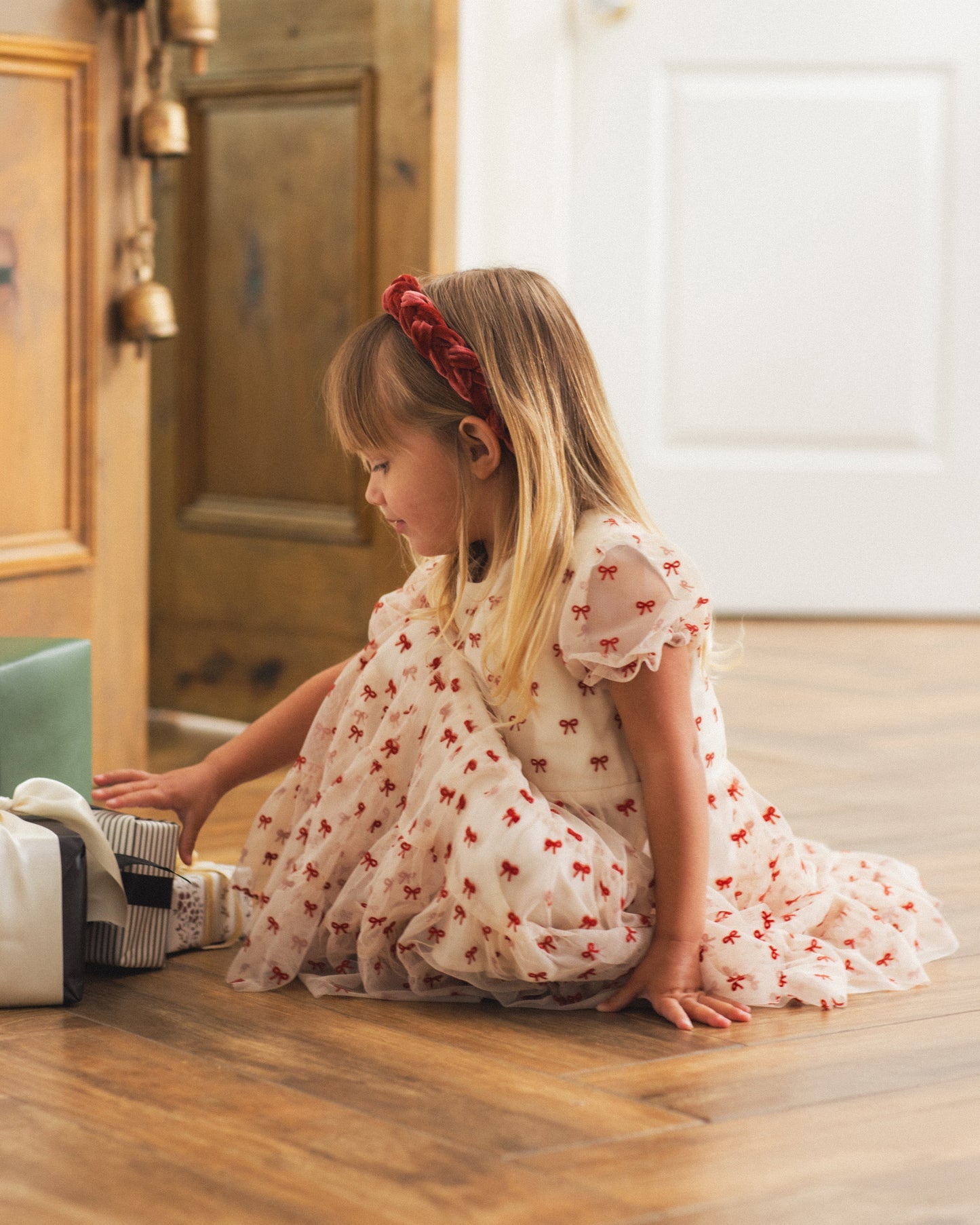 A young girl with blonde hair sits on a wooden floor wearing a white tulle dress with red bow patterns and a red bow headband, looking down at a small striped box.