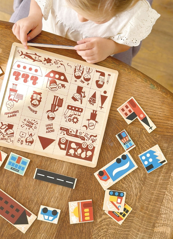 A child's hands hover above the Mentari Wooden Town Puzzle, arranging the pieces onto the illustrated wooden board on a tabletop.