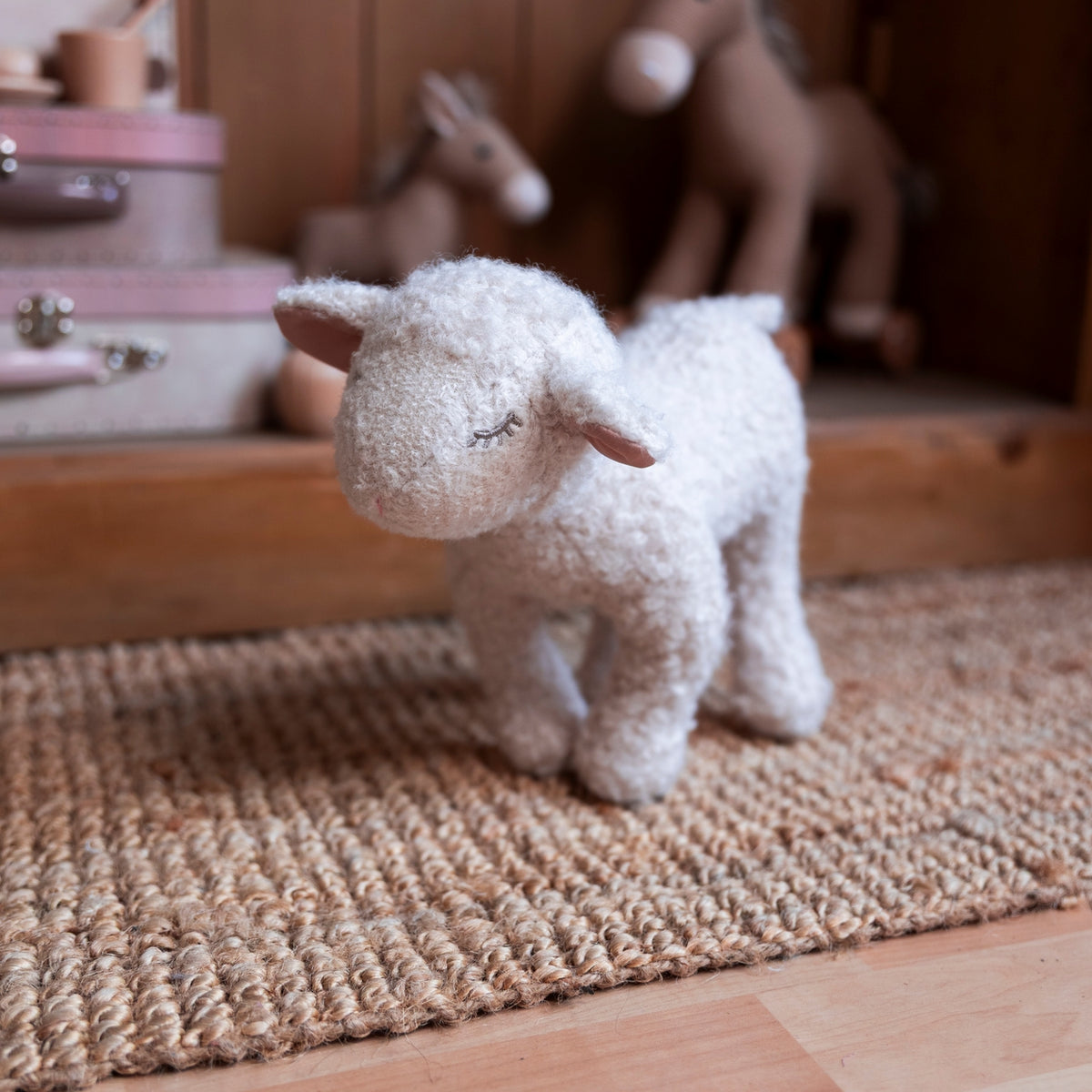 A small white plush sheep toy standing on a woven jute rug in a softly blurred nursery or playroom setting with wooden furniture and stuffed animals in the background.