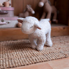 A small white plush sheep toy standing on a woven jute rug in a softly blurred nursery or playroom setting with wooden furniture and stuffed animals in the background.