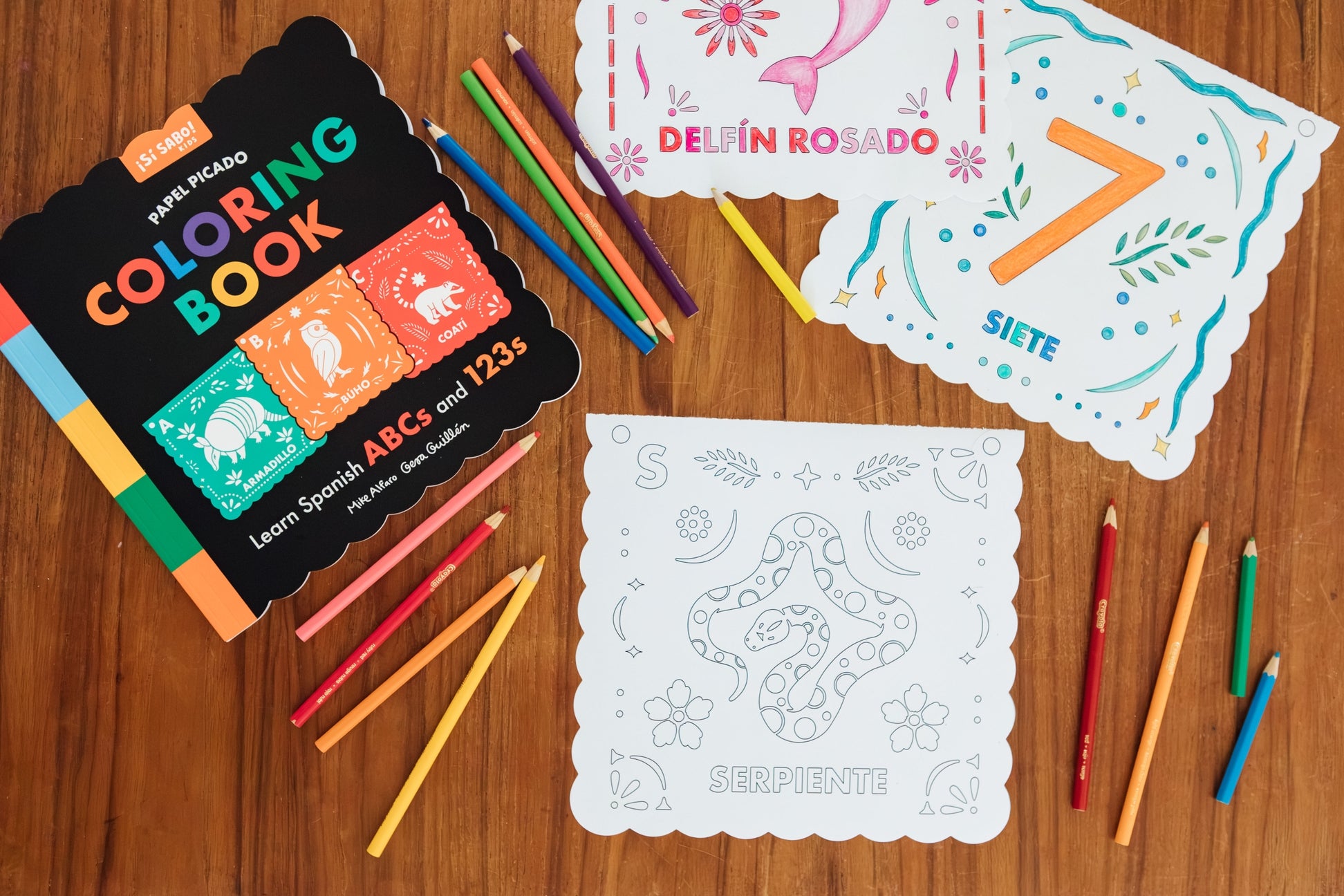 Spanish alphabet coloring book set with colored pencils and sample coloring cards arranged on a wooden surface. The set includes a black cover book labeled 'Nube Picasso' and white coloring pages with decorative Mexican-style borders.
