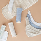 A styled flat-lay of baby muslin textiles on a warm beige background, including folded and rolled bibs and swabs in cream, light blue, and pale mint tones.