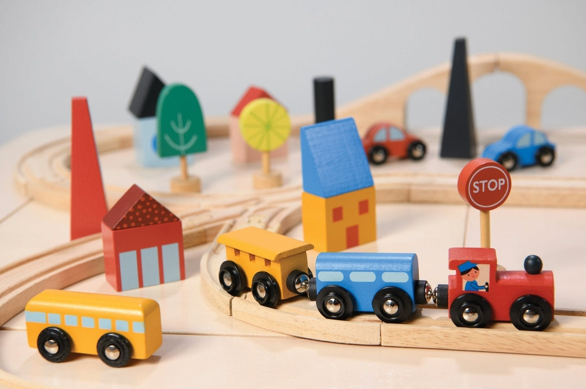 A close-up, angled view of a wooden toy train set featuring a red engine with a driver, a blue passenger car, and a yellow cargo car resting on wooden tracks. A round red "STOP" sign stands beside the track, and the background is filled with colorful wooden town elements, including a red house with a window, a yellow building, a blue bridge, and abstract green trees.