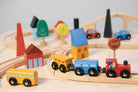 A close-up, angled view of a wooden toy train set featuring a red engine with a driver, a blue passenger car, and a yellow cargo car resting on wooden tracks. A round red "STOP" sign stands beside the track, and the background is filled with colorful wooden town elements, including a red house with a window, a yellow building, a blue bridge, and abstract green trees.