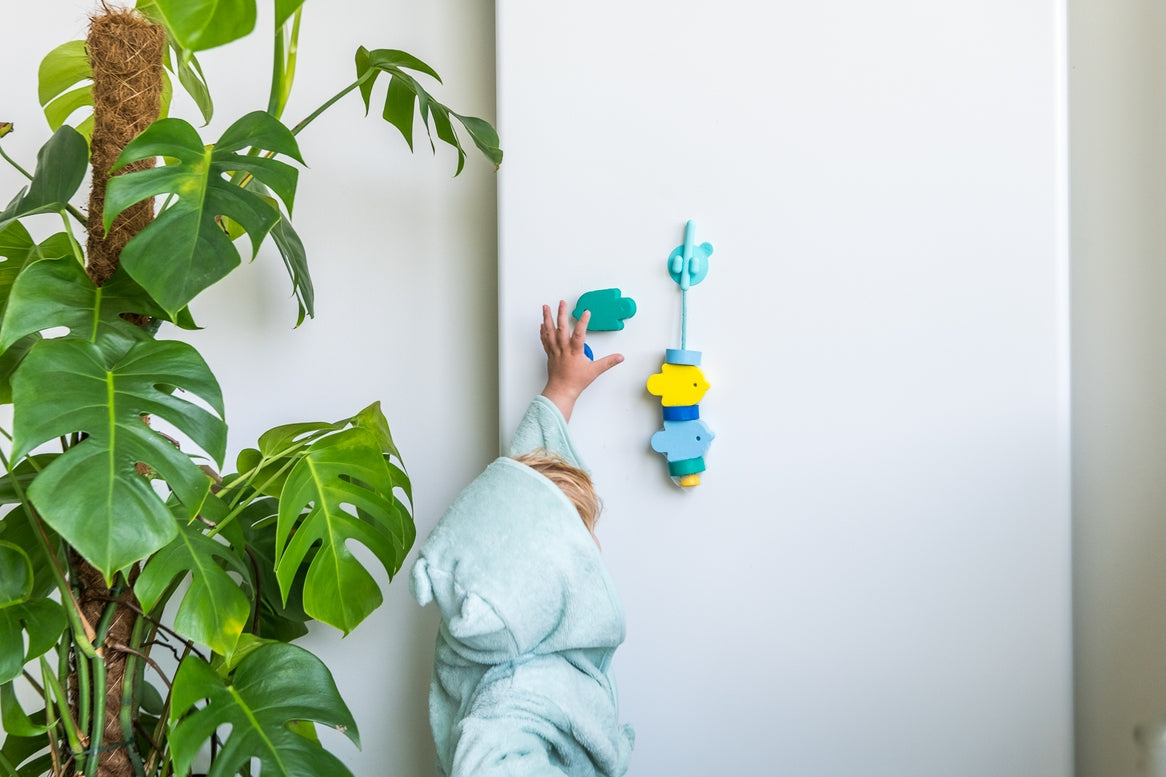 A child in a light blue hooded robe reaches for colorful wall hooks mounted on a white wall, with a large green houseplant visible on the left.