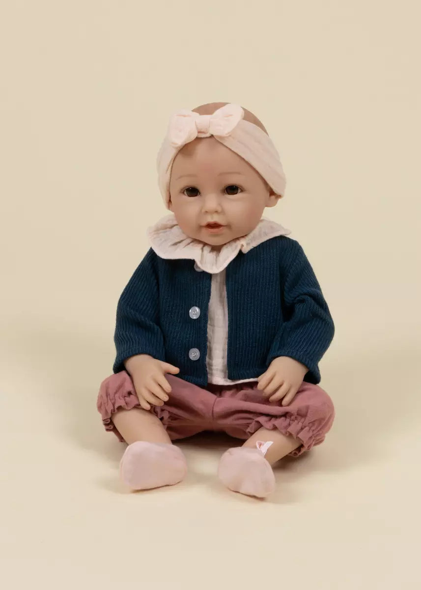 Baby doll wearing a blue cardigan, pink pants, and a pink headband on a beige background