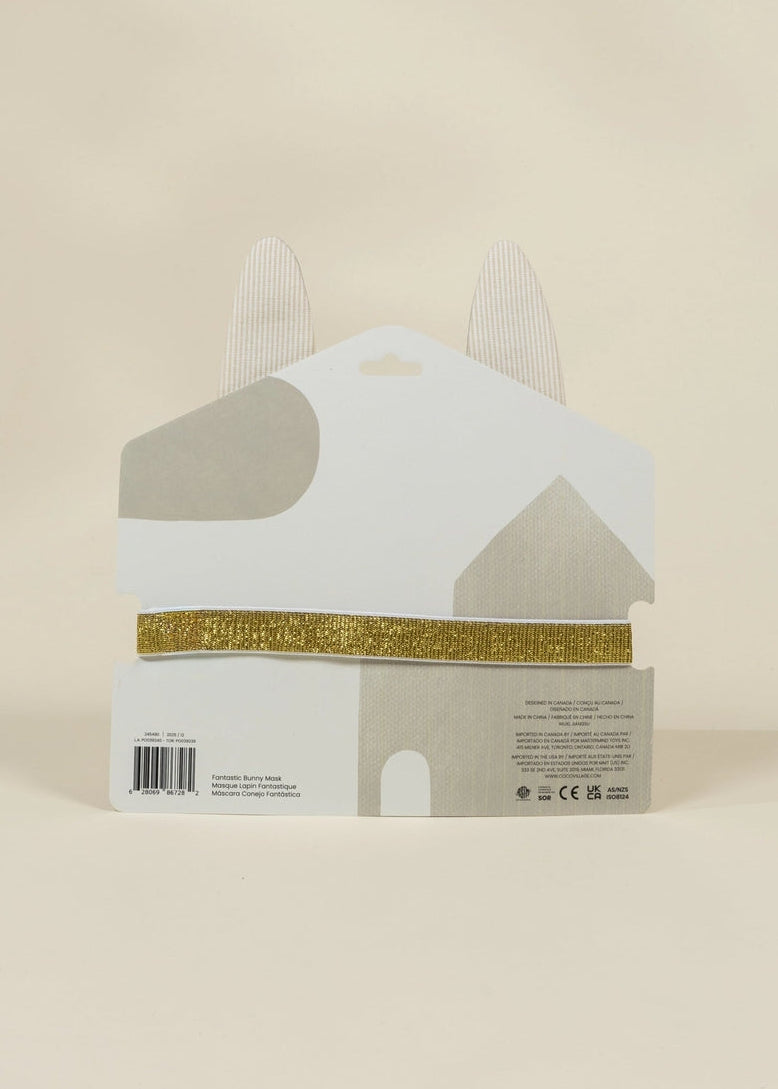 A white house-shaped product package with gray abstract shapes, a gold glitter band across the middle, and two pointed white tabs at the top, photographed against a plain warm beige background.