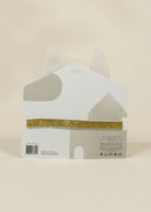 A white house-shaped product package with gray abstract shapes, a gold glitter band across the middle, and two pointed white tabs at the top, photographed against a plain warm beige background.