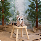 A plush Raccoon by Warmies sitting on a small wooden stool against a painted woodland backdrop with pine trees.