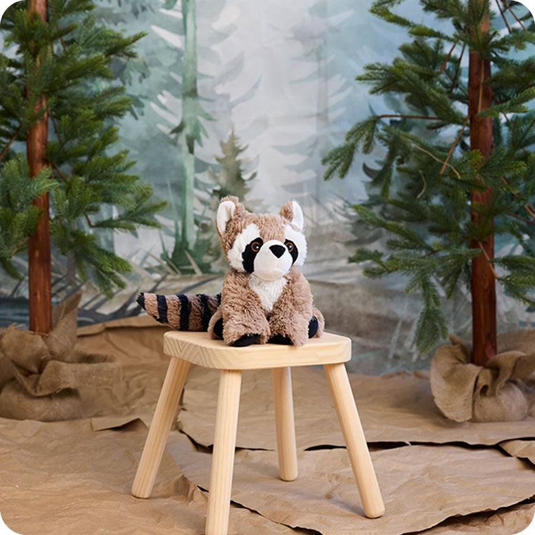 A plush Raccoon by Warmies sitting on a small wooden stool against a painted woodland backdrop with pine trees.