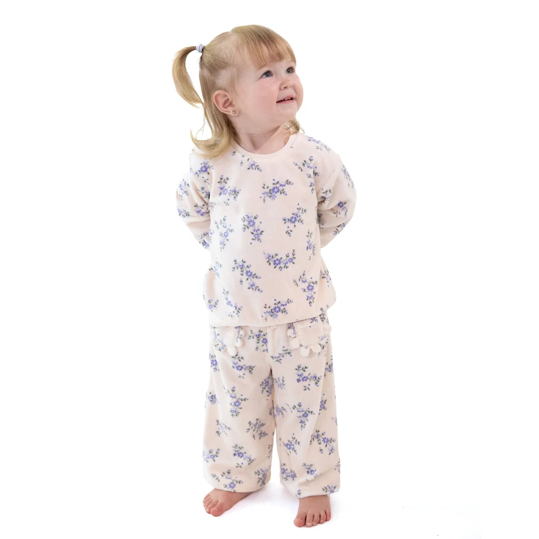 A young girl with blonde hair in a ponytail stands barefoot against a white background, wearing a two-piece pajama set with a light beige base and a blue floral pattern.