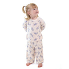 A young girl with blonde hair in a ponytail stands barefoot against a white background, wearing a two-piece pajama set with a light beige base and a blue floral pattern.