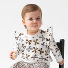A baby wearing a white long-sleeve top with brown bow and flower patterns, paired with leopard-print pants, sitting on a black chair against a light gray background.