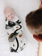 Baby swaddled in a patterned blanket with another child partially visible in the foreground.