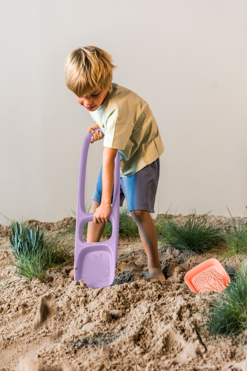 Child playing in the sand with a purple shovel and pink toy.