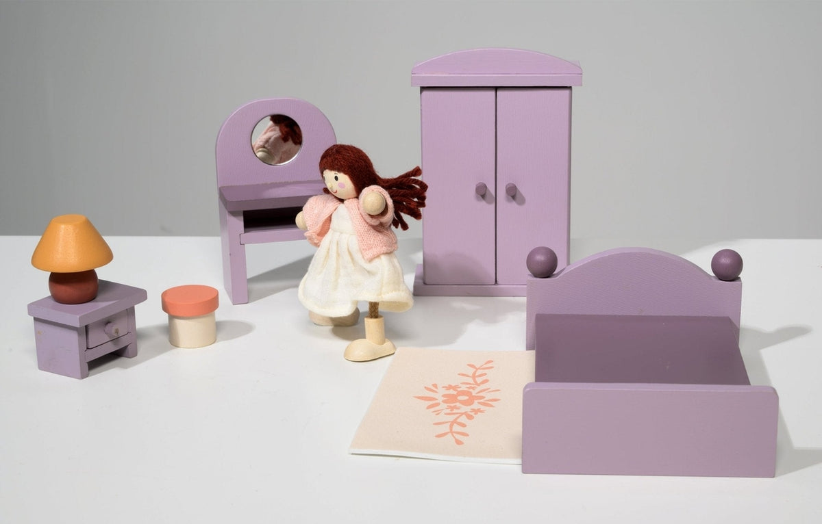 A dollhouse bedroom scene featuring a wooden doll in a pale dress standing near the center, positioned in front of a purple double bed. In the background, a matching purple wardrobe stands beside a dressing table with an oval mirror and a stool, while a bedside table with an orange-based lamp topped with a yellow shade sits on the left.