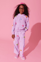 A young girl with curly hair and glasses wearing a light purple/lavender matching lounge set with colorful flower appliqués, standing against a solid pink studio background.