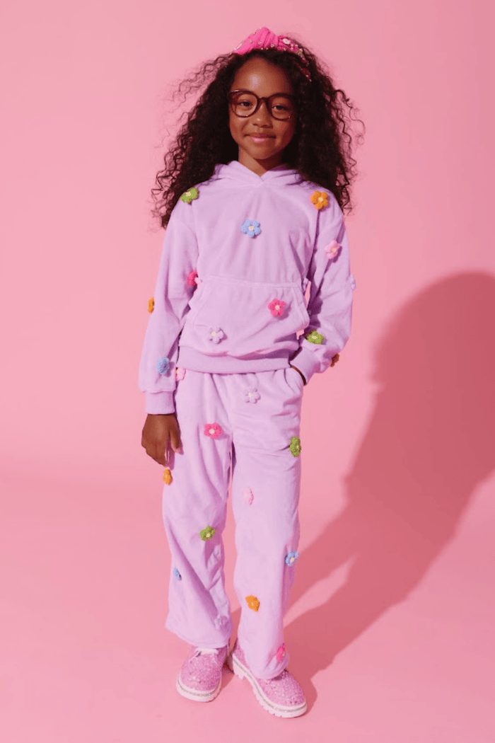 A young girl with curly hair and glasses wearing a light purple/lavender matching lounge set with colorful flower appliqués, standing against a solid pink studio background.