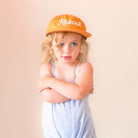 A young child with blonde curly hair wearing a light blue and white gingham romper and an orange baseball cap with 'Rascal' embroidered in white, posed against a solid beige background.